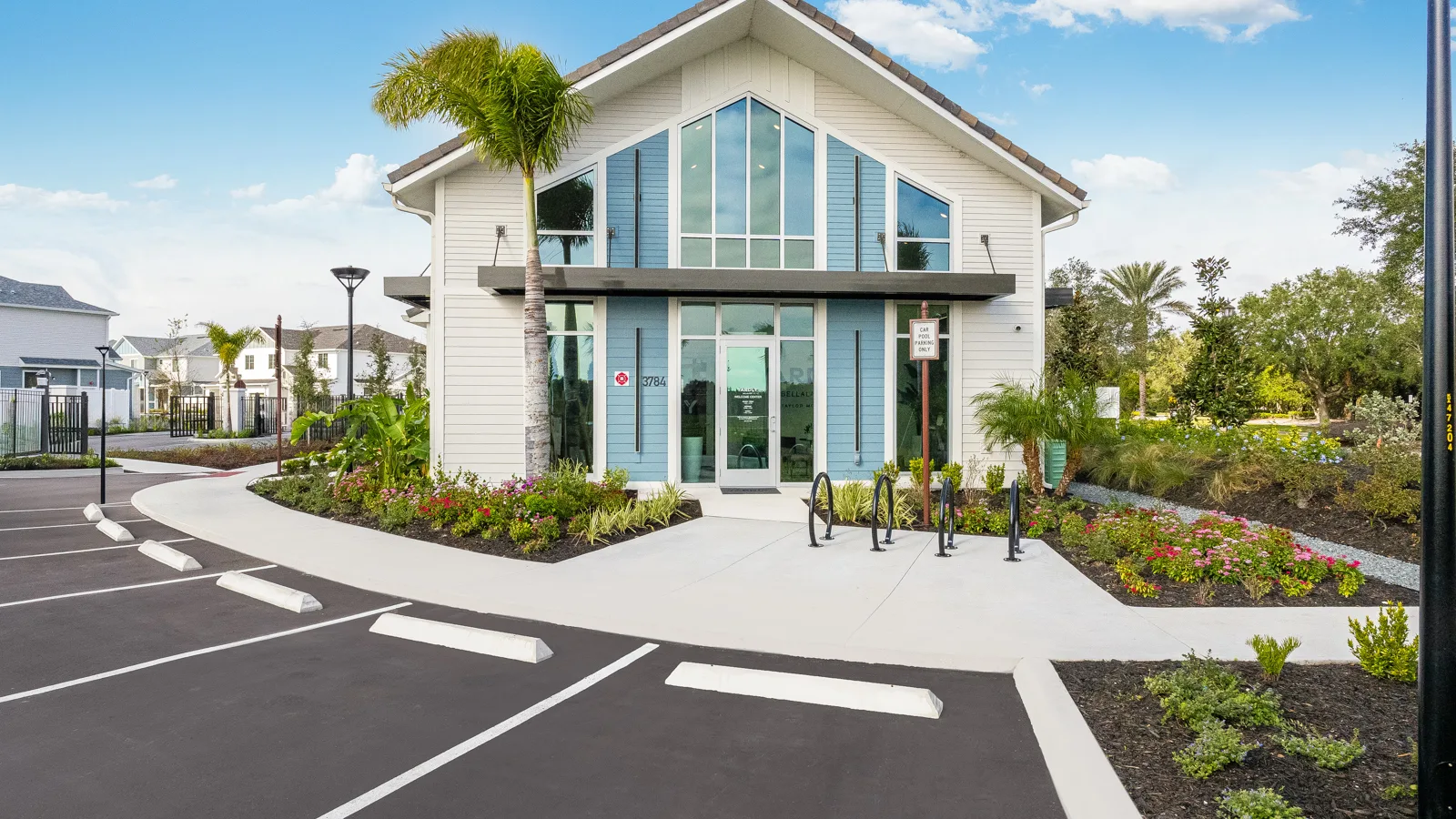 Exterior of leasing office and fitness center with a bike rack, plenty of parking and luscious green tropical foliage. Community sidewalk facing pond with green grass and trees. Apartments in Orlando with a view of beautiful landscape. New pet-friendly apartment homes for rent with private fenced backyards in Kissimmee, FL, near Orlando, FL with pickle ball, dog park, gym, and gated community. *No breed or weight restrictions. Yardly Bellalago