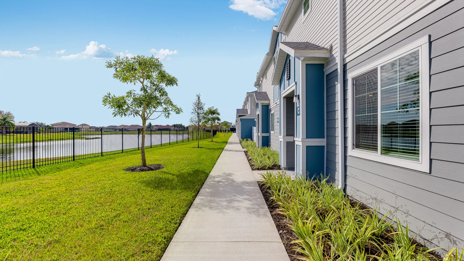 Community sidewalk facing pond with green grass and trees. Apartments in Orlando with a view of beautiful landscape. New pet-friendly apartment homes for rent with private fenced backyards in Kissimmee, FL, near Orlando, FL with pickle ball, dog park, gym, and gated community. *No breed or weight restrictions. Yardly Bellalago